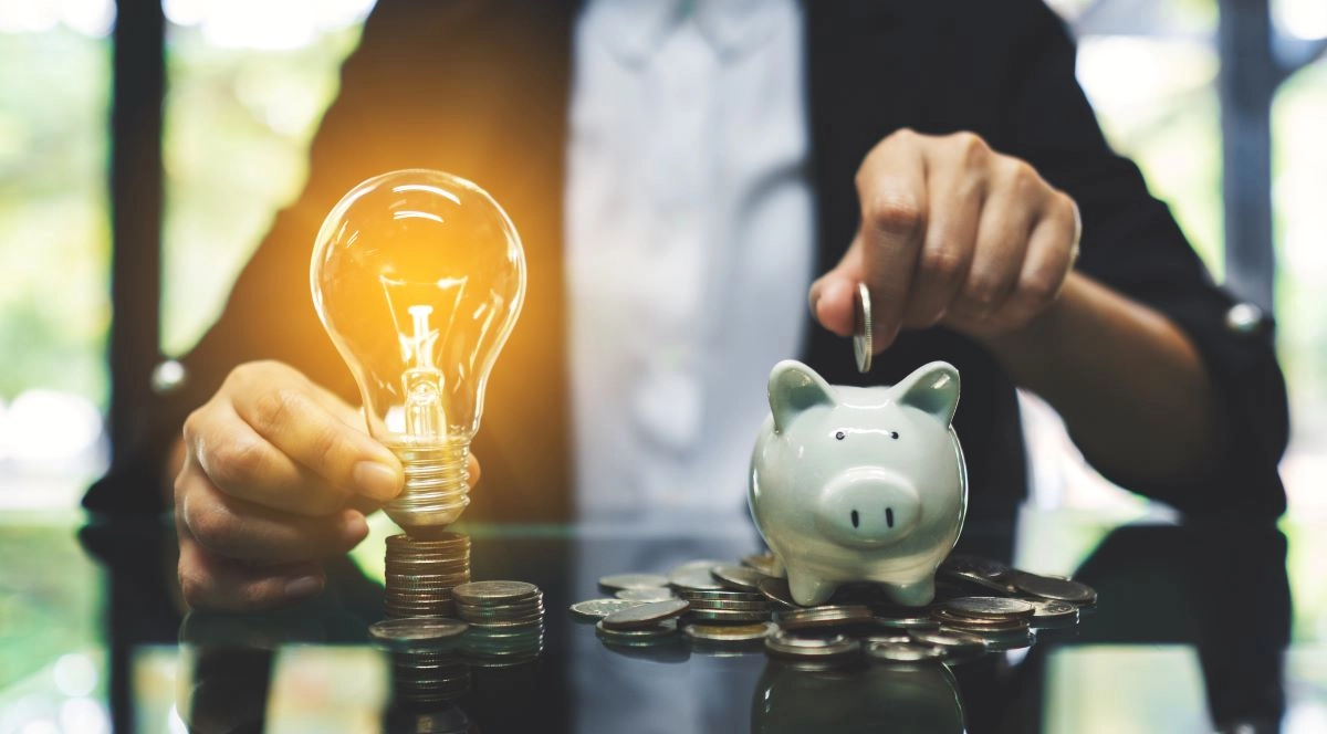 Person placing coin into piggy bank next to glowing light bulb, representing energy savings and cost efficiency